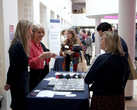 Two Caucasian females standing behind a desk with leaflets on speak to a Caucasian male and female.